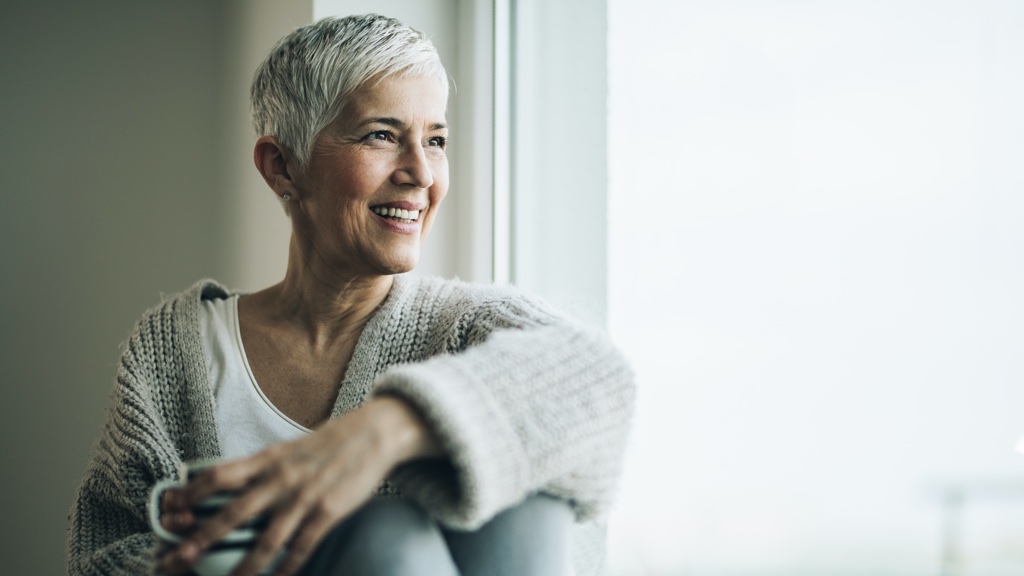 Happy mature woman with coffee cup relaxing by the window. Who We Serve